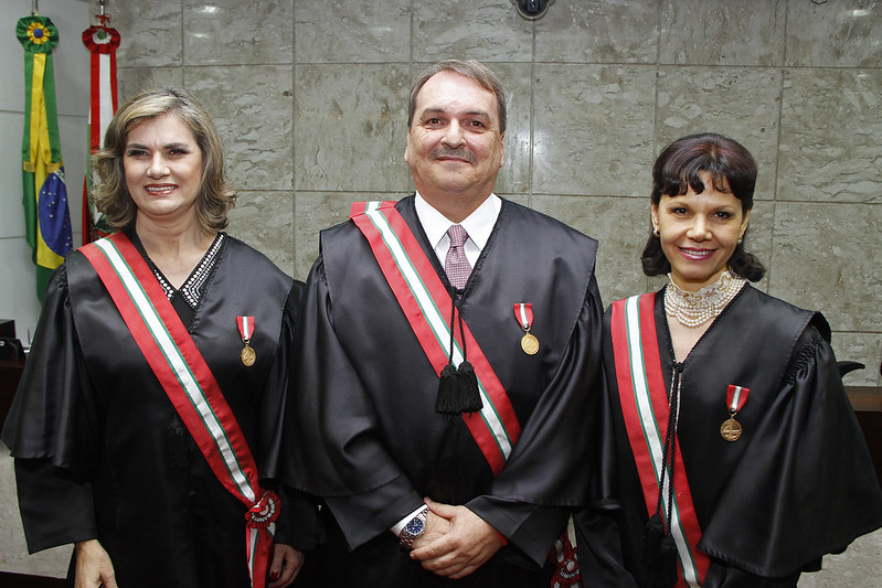 Foto de um homem no centro de duas mulheres. Os três estão usando toga e sorrindo