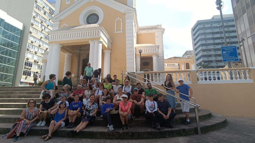 Foto com um grupo de pessoas sentados na escadaria em frente a uma igreja