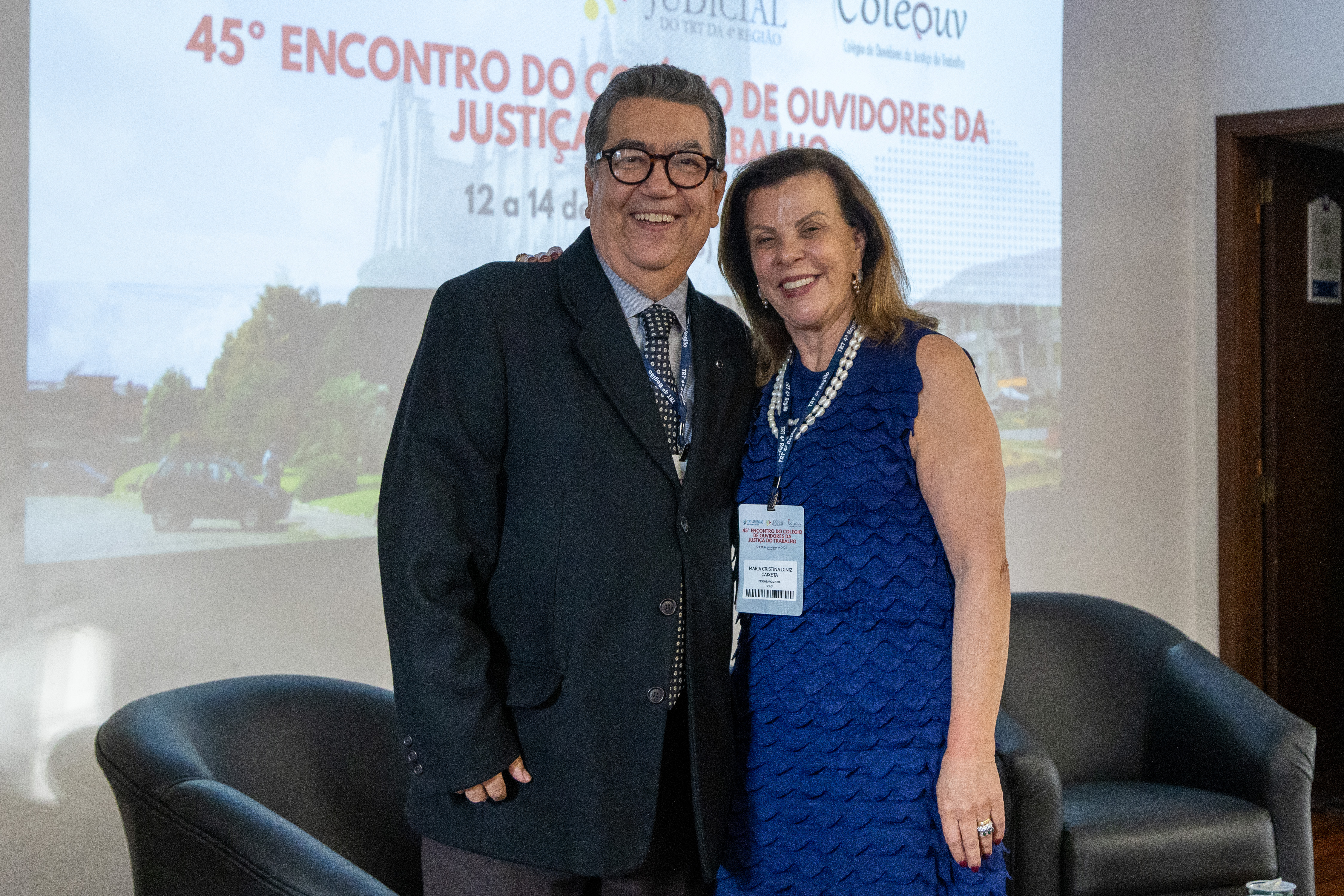 Homem de terno e mulher de vestido azul posam para foto sorridentes, à frente de um telão e de duas poltronas pretas
