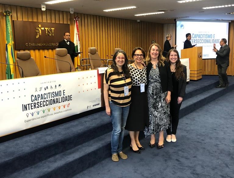 Quatro mulheres em pé posam para foto, sorridentes, em um amplo auditório