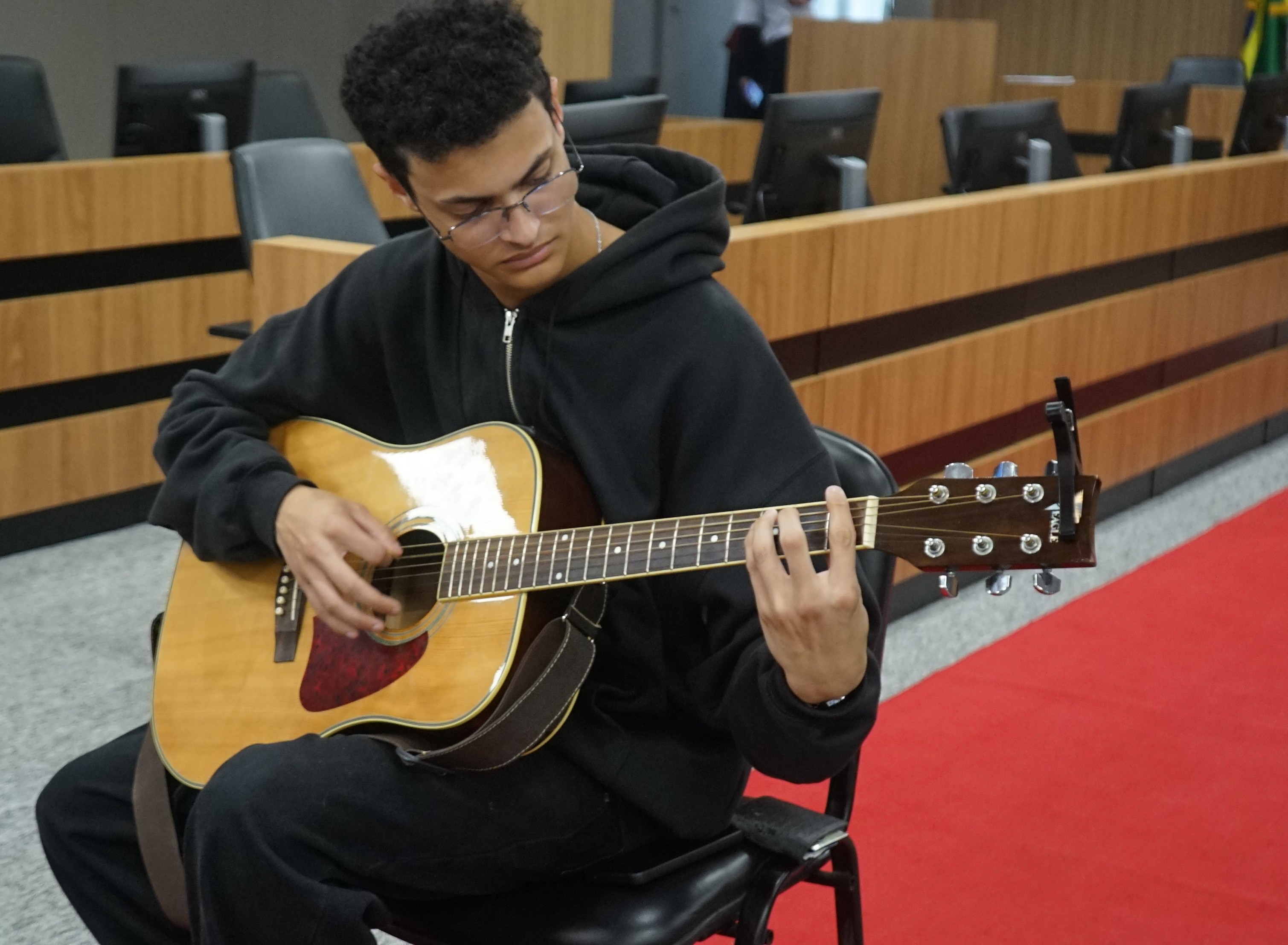 Jovem mestiço, de cabelos negros, usando óculos e tocando violão