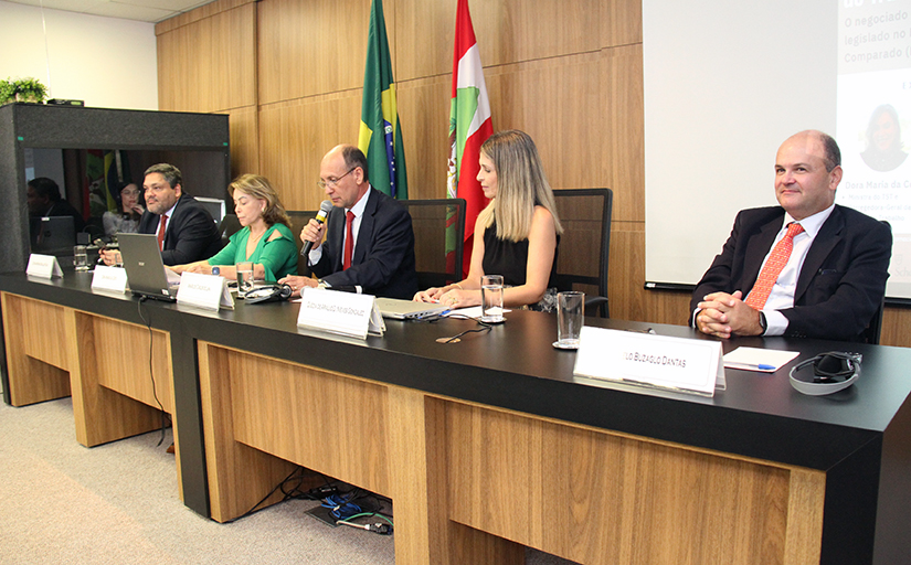 Cinco pessoas estão sentadas em uma mesa, em um auditório, sendo três homens e duas mulheres. Ao centro, o presidente do TRT-SC, Amarildo Carlos de Lima, fala ao microfone.