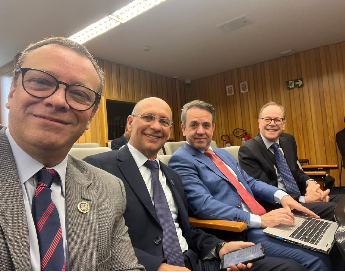 Foto no estilo selfie mostrando quatro homens sentados, de terno, sorridentes para a câmera. Eles estão em um auditório e um deles tem um notebook no colo.