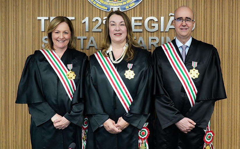 A imagem mostra duas magistradas e um magistrado posando lado a lado, usando togas pretas e faixas nas cores verde, branca e vermelha, com medalhas douradas presas ao peito