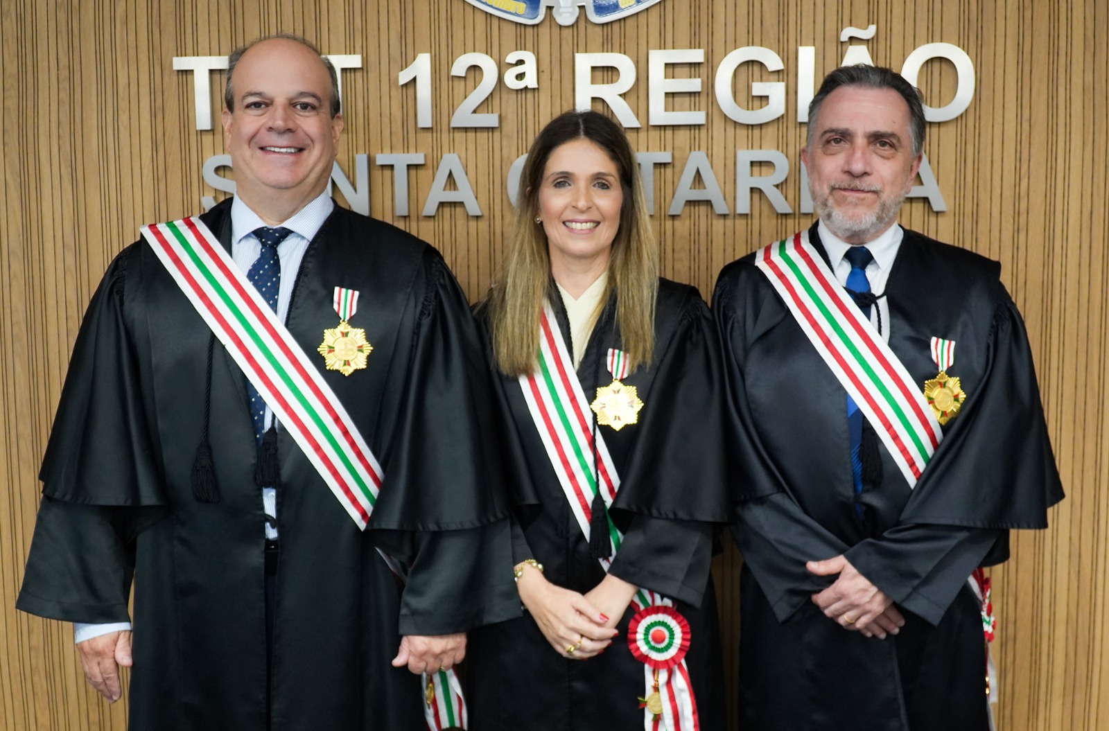 Três pessoas vestidas com toga, faixa verde, vermelhe e branco em torno da toga e uma medalha fixada nela.