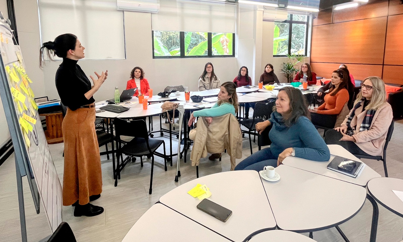 Foto de 12 mulheres reunidas em uma sala, a maioria delas está sentada de frente para uma mesa ou virada para a única mulher em pé, que gesticula com os braços ao lado de um quadro cheio de notas adesivas amarelas