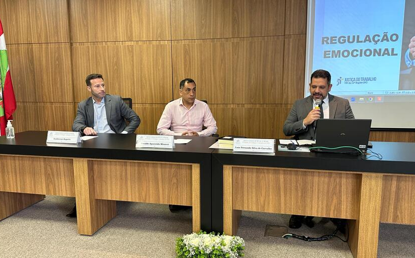 Fotografia de três homens vestidos formalmente sentados atrás de uma bancada de madeira. Um deles tem um notebook à frente e fala segurando um microfone