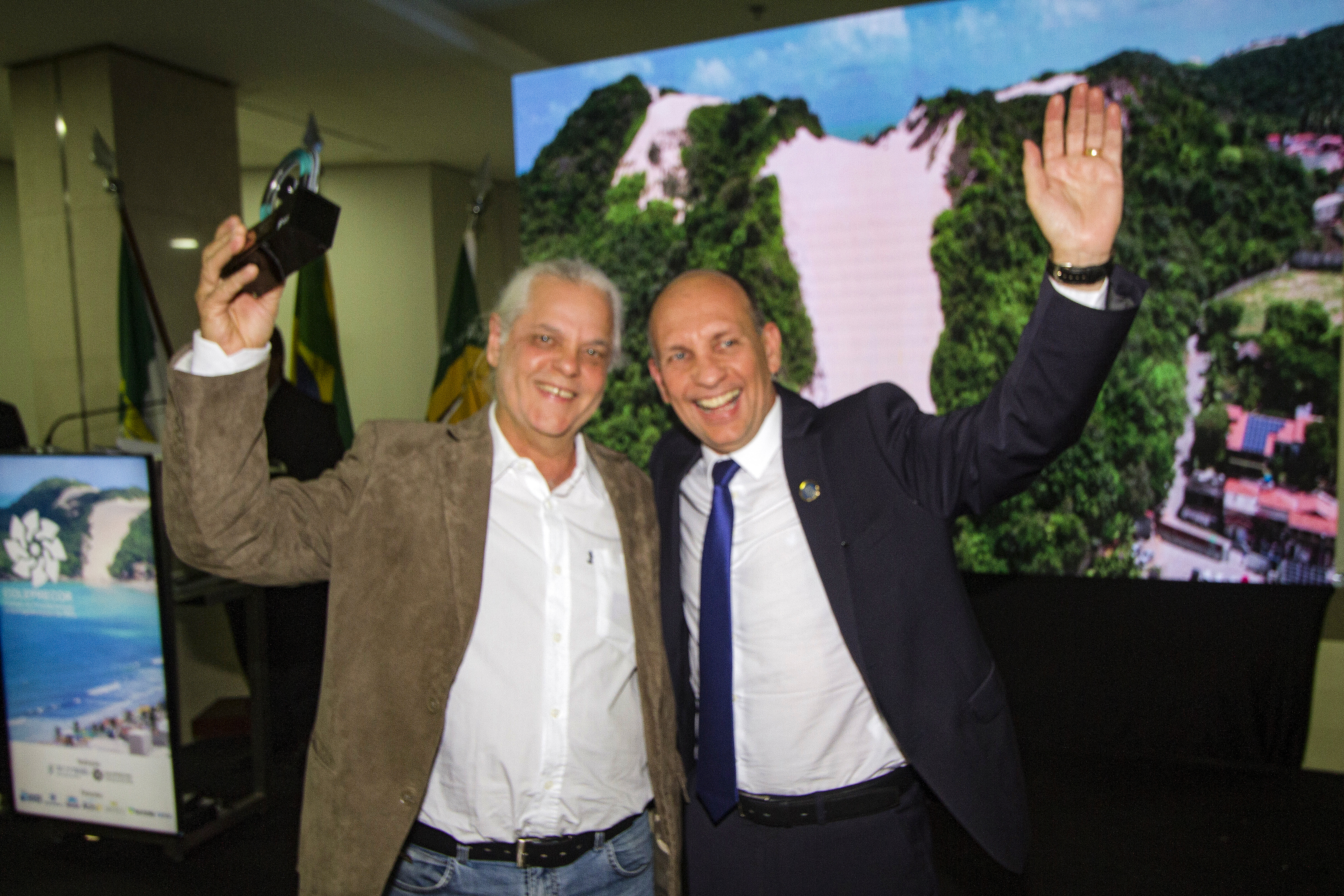 Foto de dois homens em gesto de comemoração sorrindo, se abraçando com um braço e erguendo o outro. O homem à esquerda é branco, tem o cabelo grisalho curto, usa um paletó marrom e segura na mão livre um troféu. O outro homem é branco, calvo e usa terno azul escuro com gravata azul. Ao fundo um ambiente escuro com um telão luminoso mostrando um cenário natural