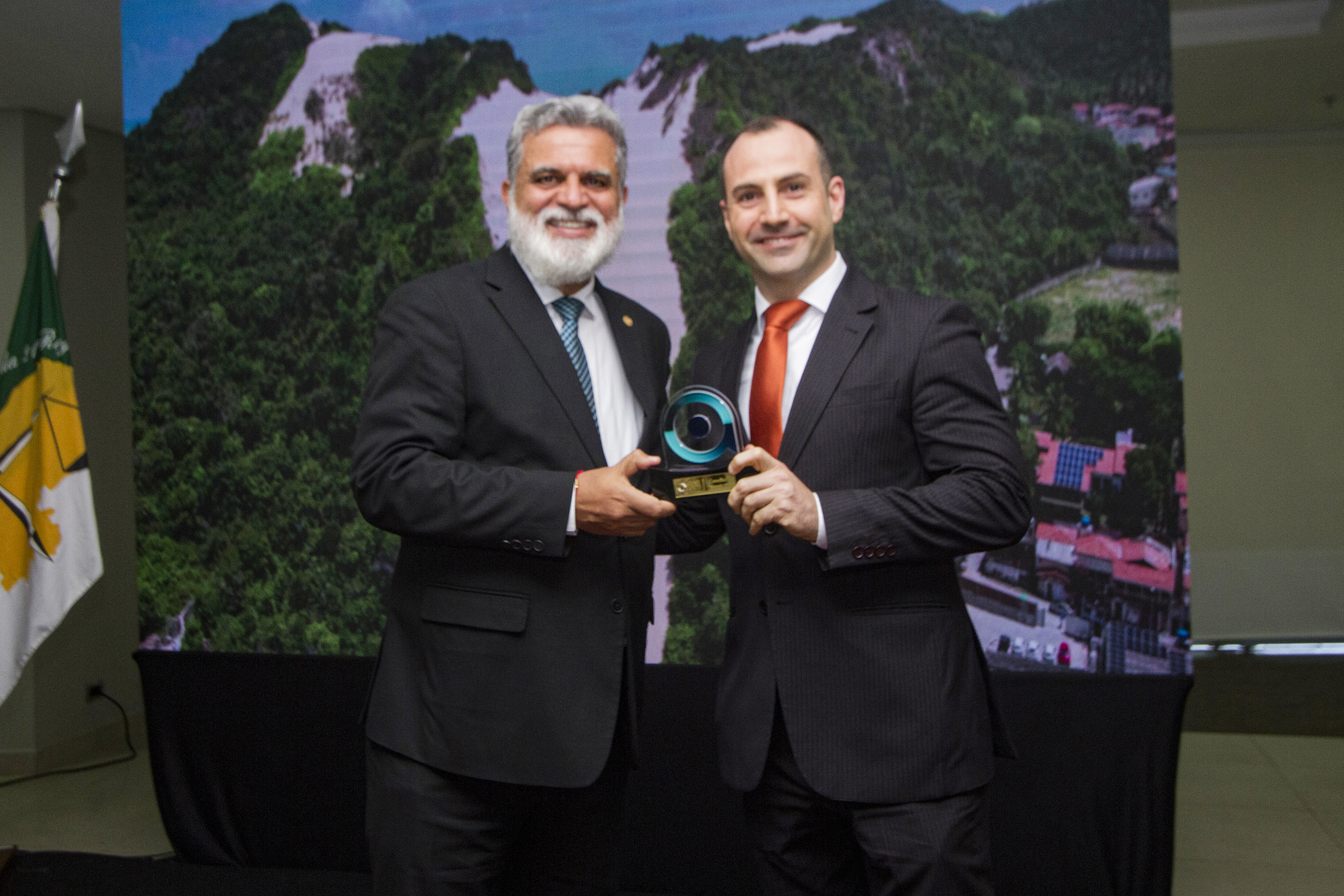 Foto posada de dois homens, cada um segurando com uma mão o mesmo troféu. O homem à esquerda é branco, tem cabelo e barba grisalha e usa terno escuro com grava azul listrada, ao seu lado um homem branco, de cabelo escuro, calvo, usando terno escuro e gravata alaranjada. Ao fundo um telão com o mesmo cenário da foto anterior 