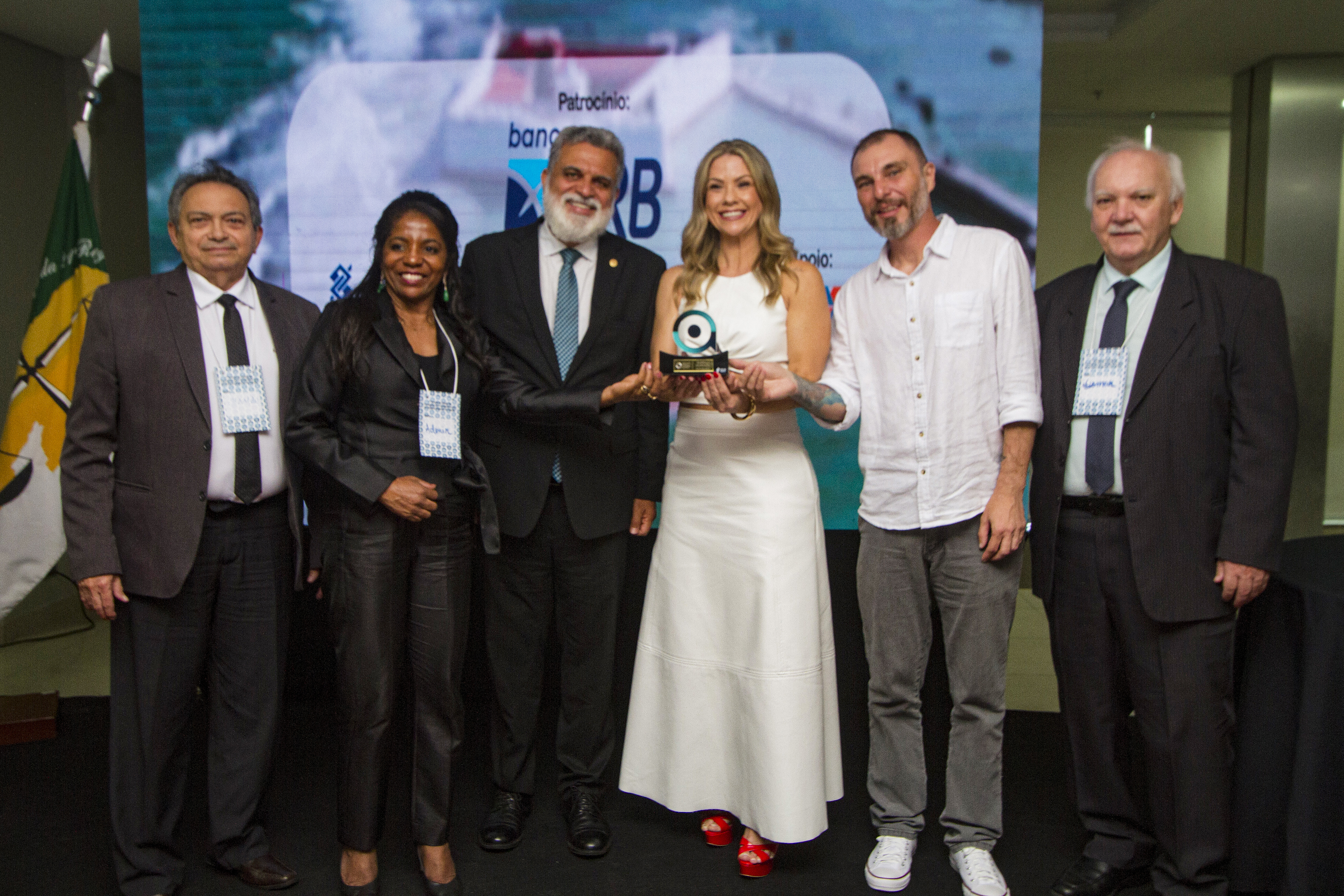 Foto de seis pessoas, três segurando o mesmo troféu. São duas mulheres e quatro homens. Está presente na imagem o homem de barba grisalha e, o quinto da esquerda pra direita, um homem de cabelo curto escuro e braba grisalha vestindo camisa de botão branca e com tatuagens à mostra no braço