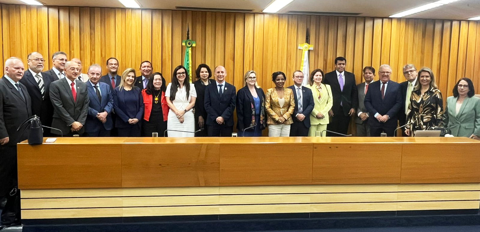 Um grupo de 23 pessoas vestidas formalmente, a maioria homens de terno, reúnem-se para uma foto. Eles estão atrás de uma longa bancada de madeira, tendo ao fundo um painel de madeira em tom quase dourado.