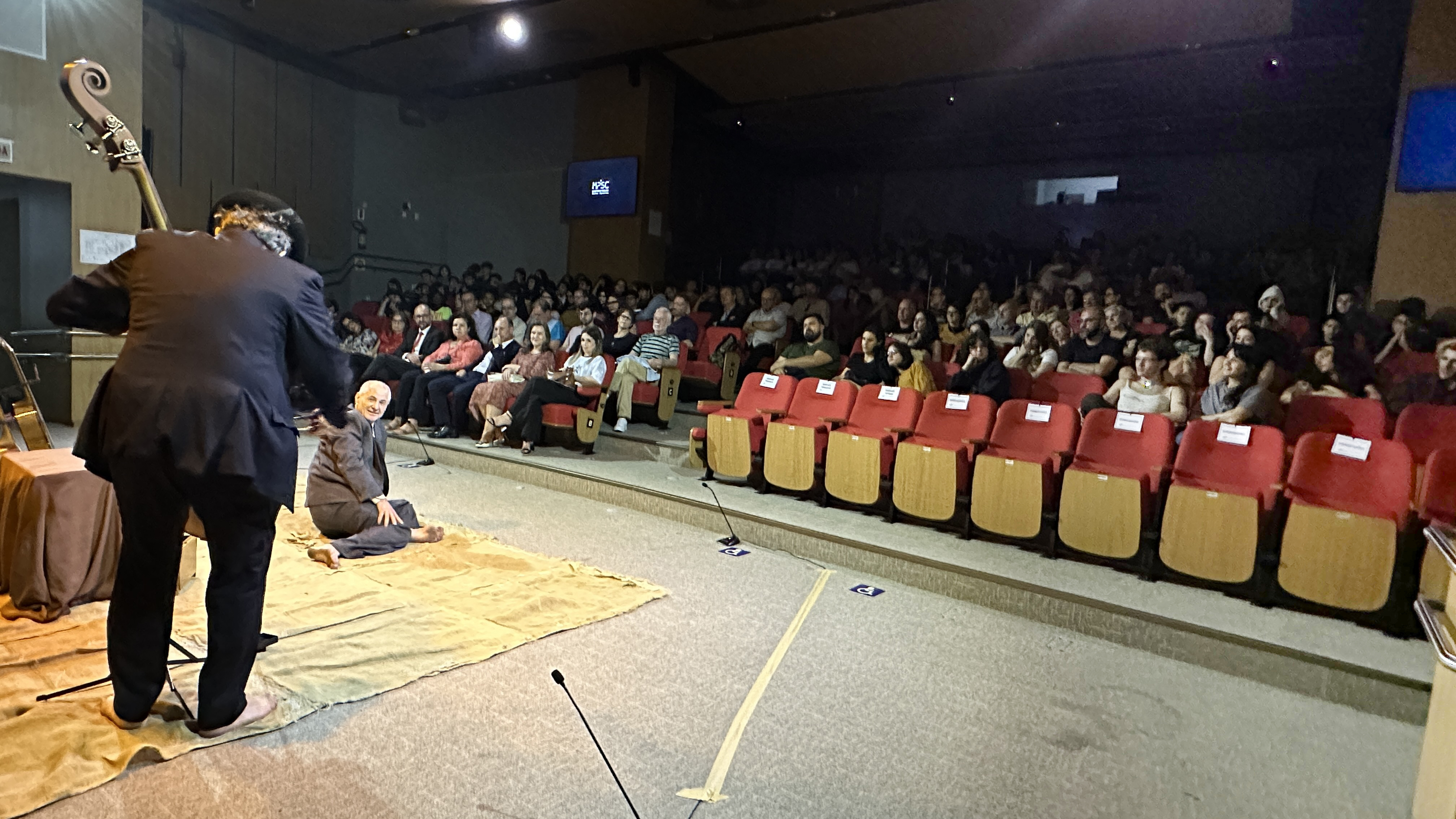 Foto de um auditório escuro cheio de pessoas, na parte iluminada estão dois homens, um sentado de lado no chão e outro de costas para a câmera, parece segurar algo que aparenta ser um contrabaixo acústico, é possível ver apenas a cabeça do instrumento, o resto está obstruído pelo corpo do homem de chapéu