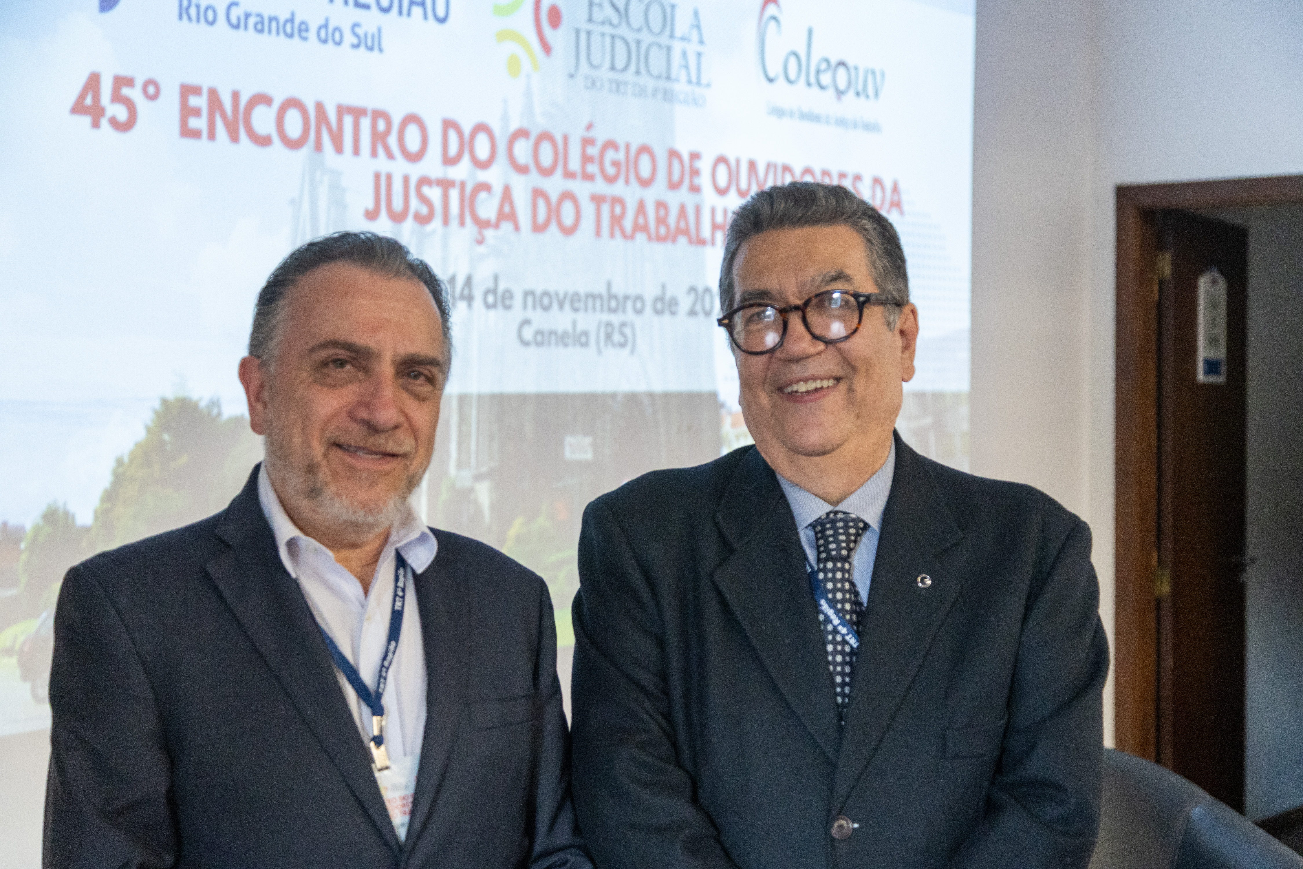 Dois homens de terno escuro posam para a foto à frente de um telão