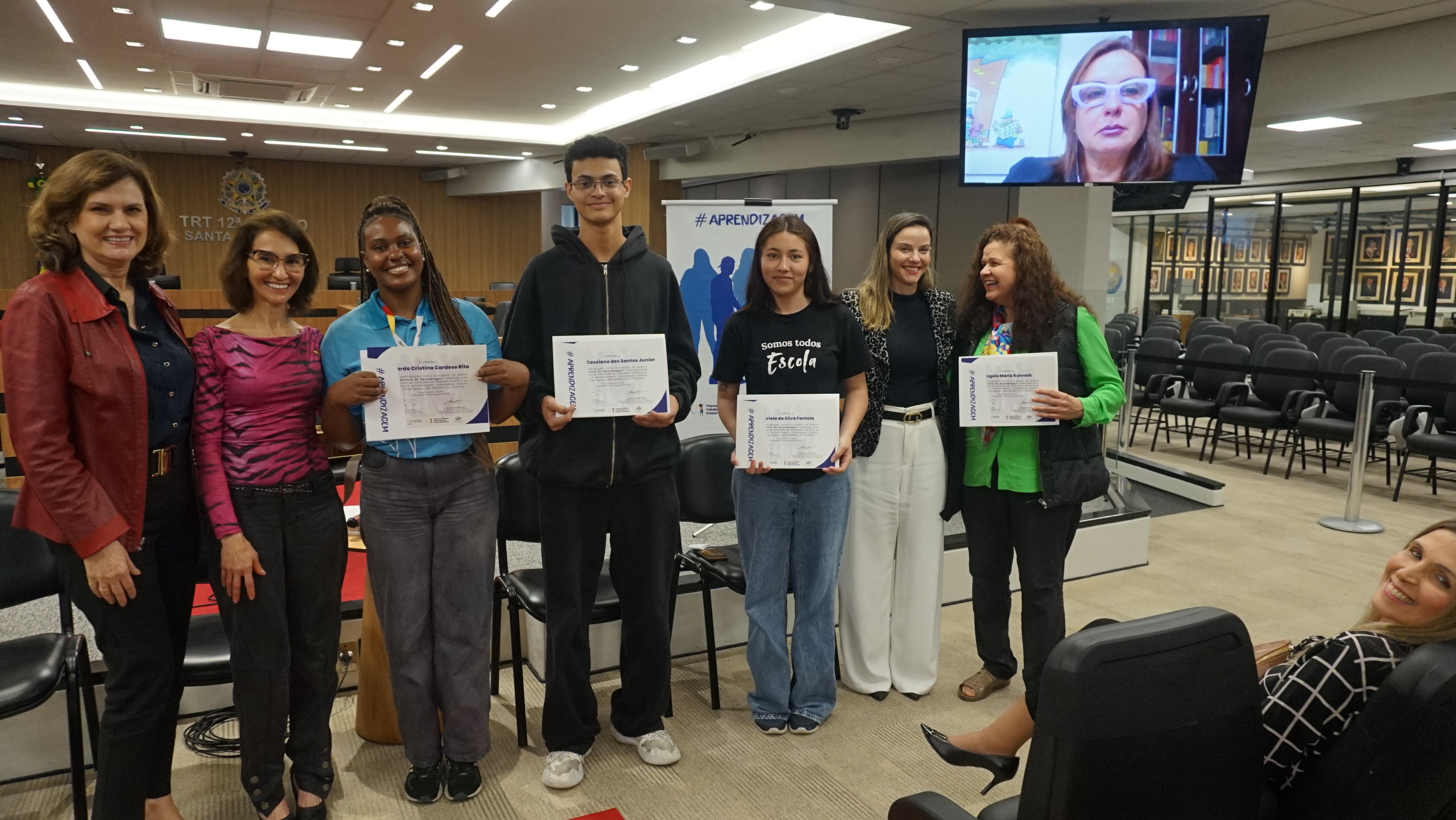 Três jovens e quatro mulheres adultas posam para foto. Os jovens e uma das mulheres seguram um certificado, No alto, um telão mostra outra mulher adulta
