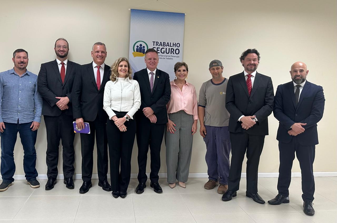 Duas mulheres e sete homens em pé posam para a foto, lado a lado. Atrás, um banner escrito Trabalho Seguro