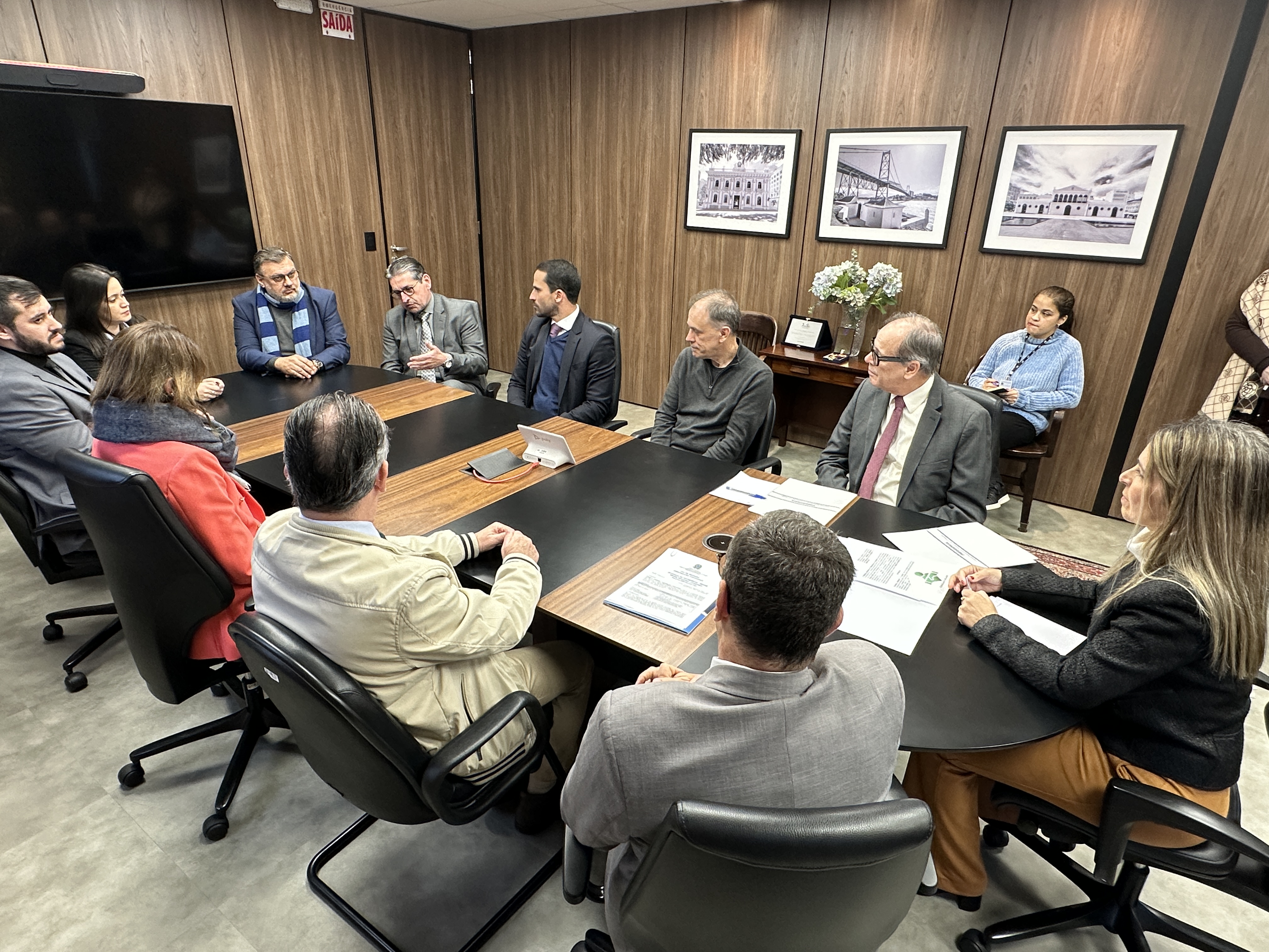Reunião com 12 pessoas em torno de uma mesa, algumas com documentos em mãos; ambiente formal e profissional.