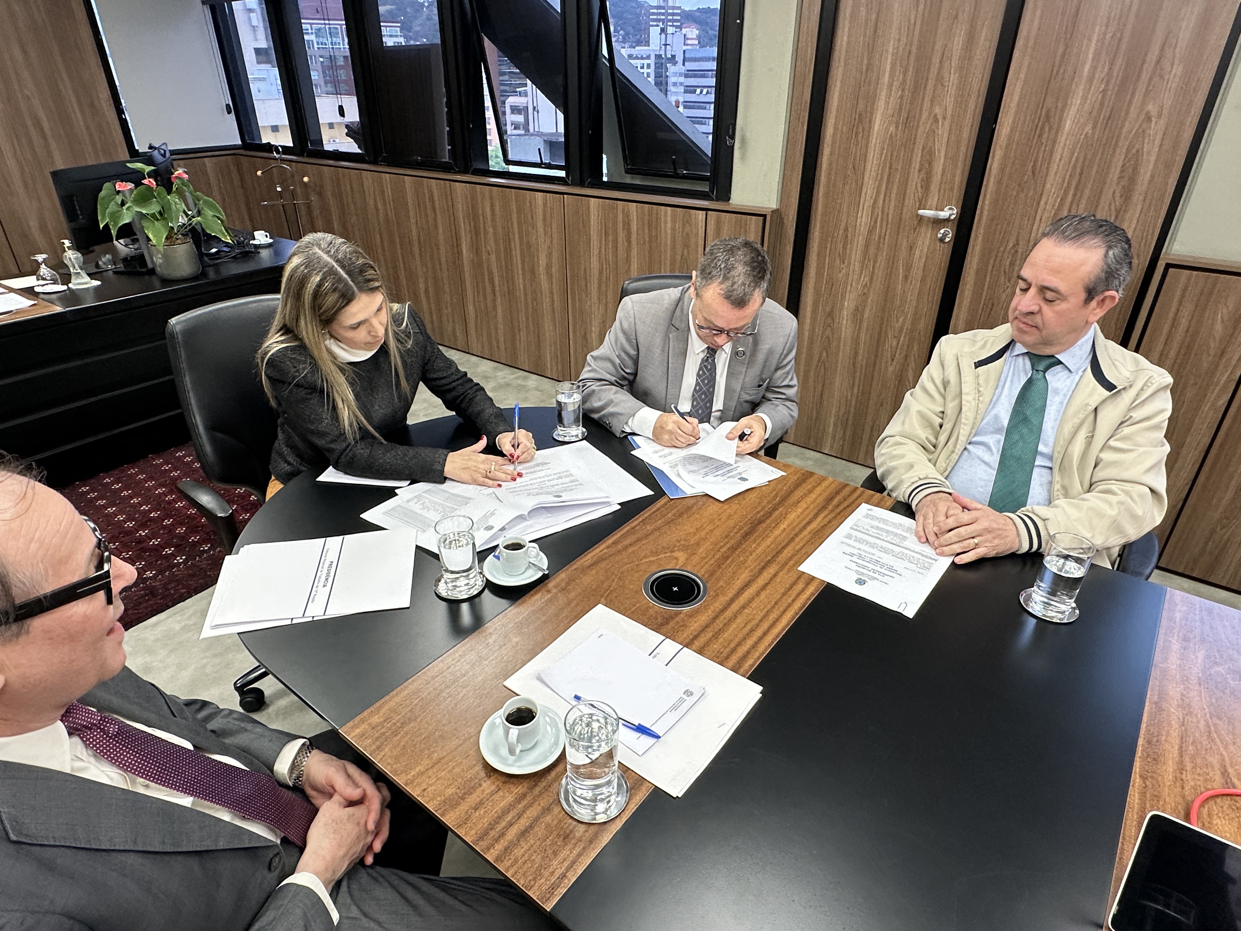 Quatro pessoas em reunião formal; duas assinam documentos enquanto as outras observam. Há papéis, café e água sobre a mesa.