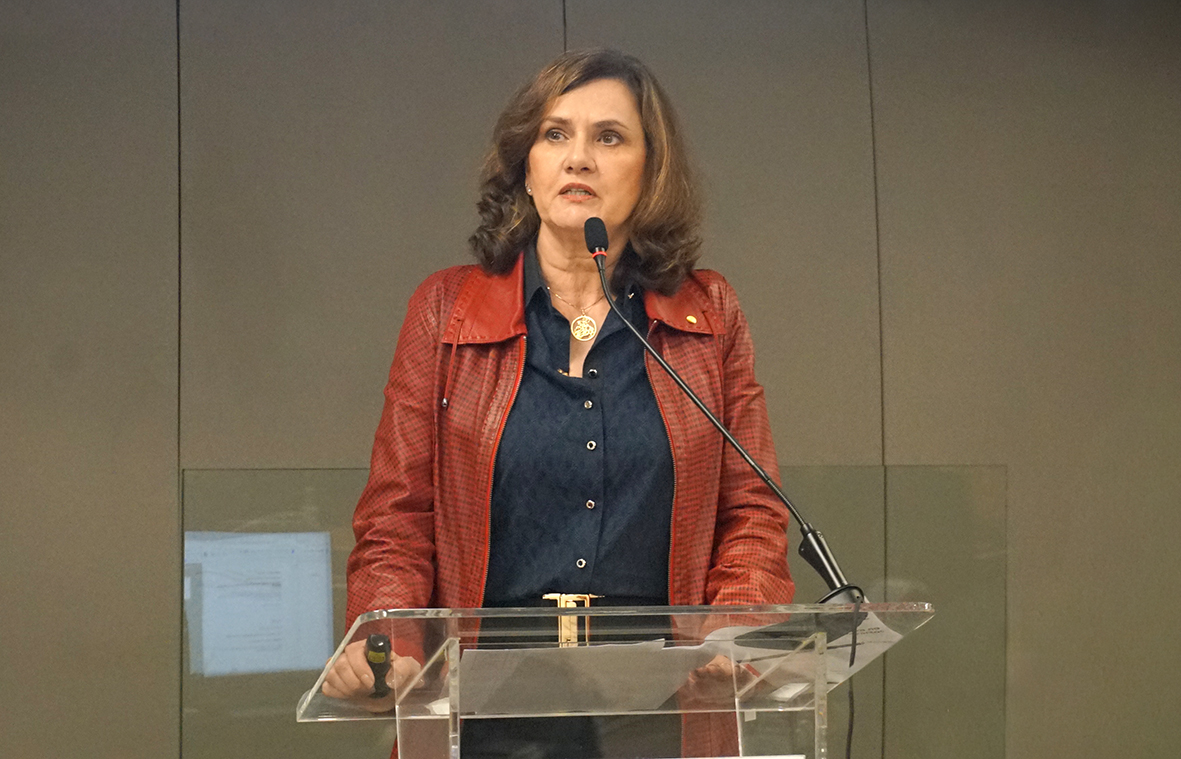 Mulher de cabelos castanhos, vestindo blusa preta e casaco de couro vermelho, fala ao microfone
