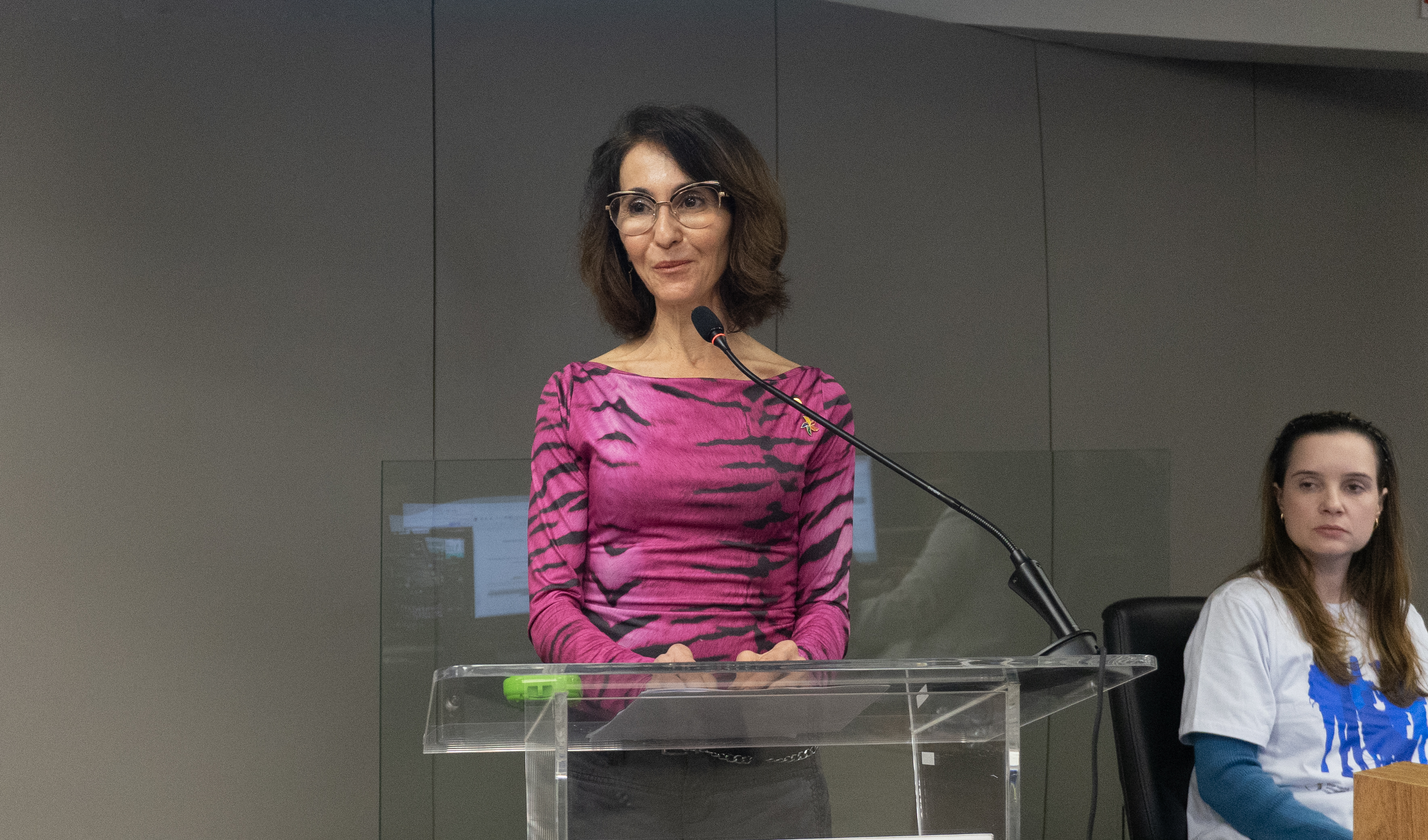Mulher de cabelos castanhos, usando óculos e vestindo uma blusa rosa, fala ao microfone