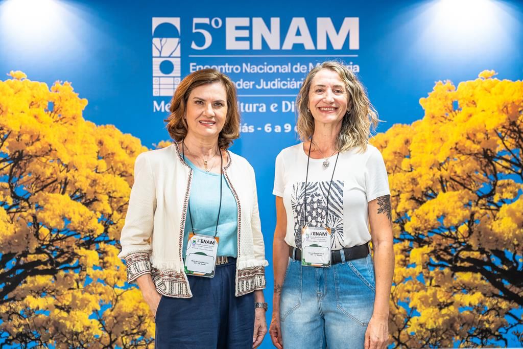 Duas mulheres em pé, em frente a um painel com fundo azul e árvores amarelas, ambas vestindo roupas casuais