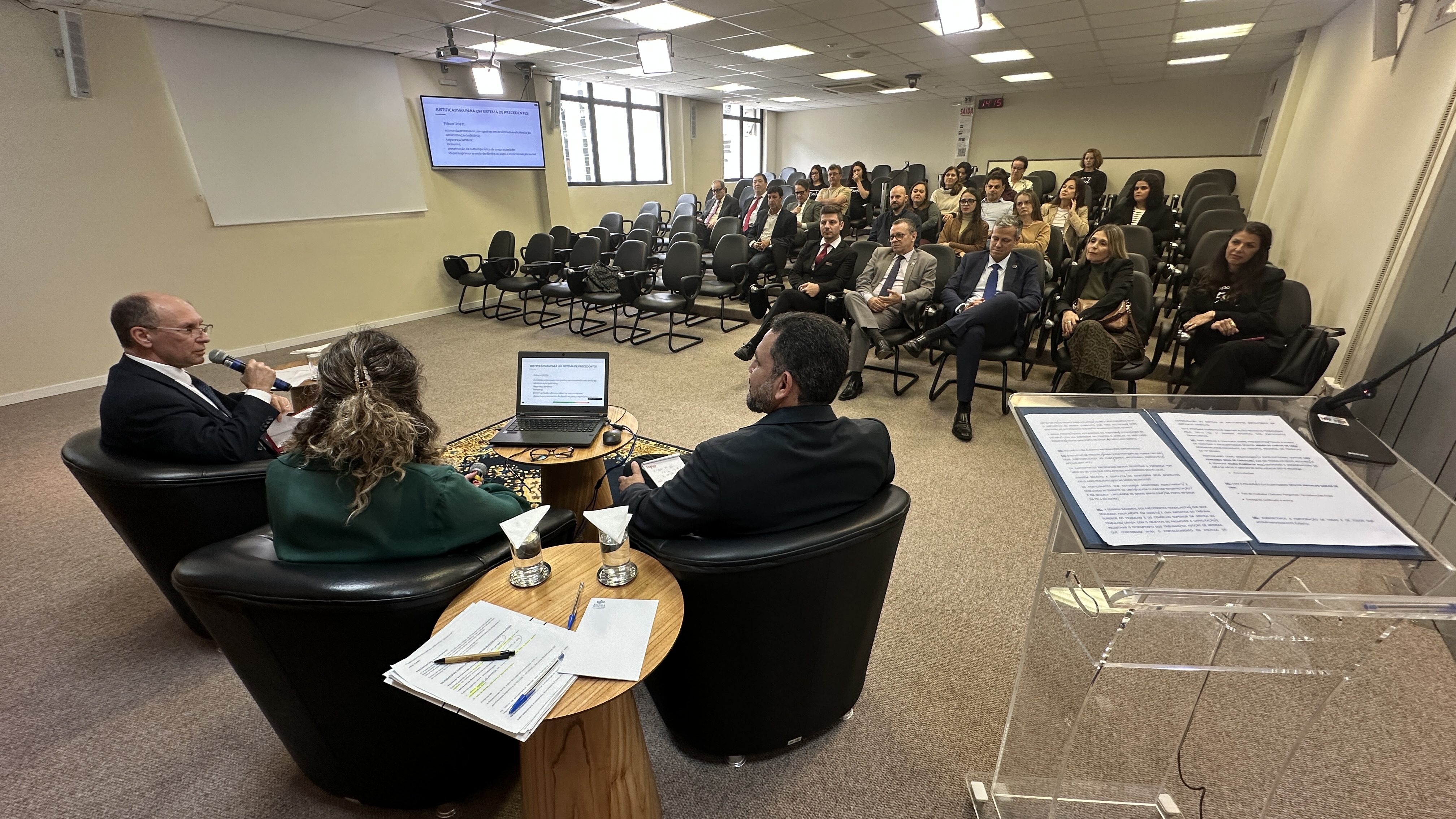 Foto de um auditório com três pessoas sentadas no palco e uma plateia assistindo