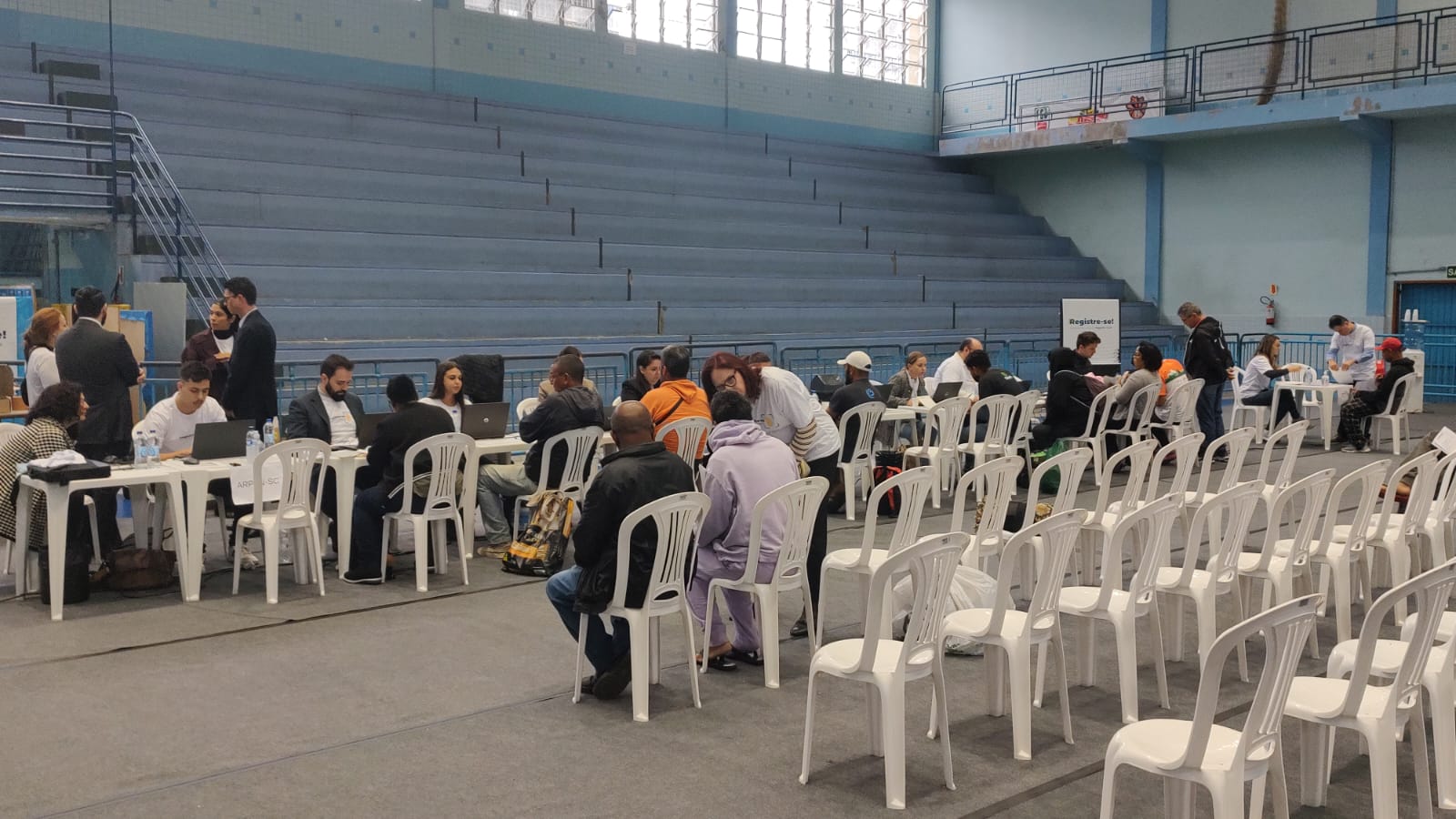 Em um ginásio estão dispostas várias mesas de atendimento ao público, com cadeiras para quem chega e aguarda. Ao fundo, equipes estão com computadores e atendem pessoas.
