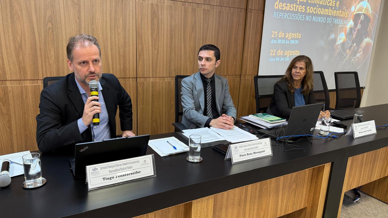 Dois homens e uma mulher vestida formalmente em uma mesa de auditório