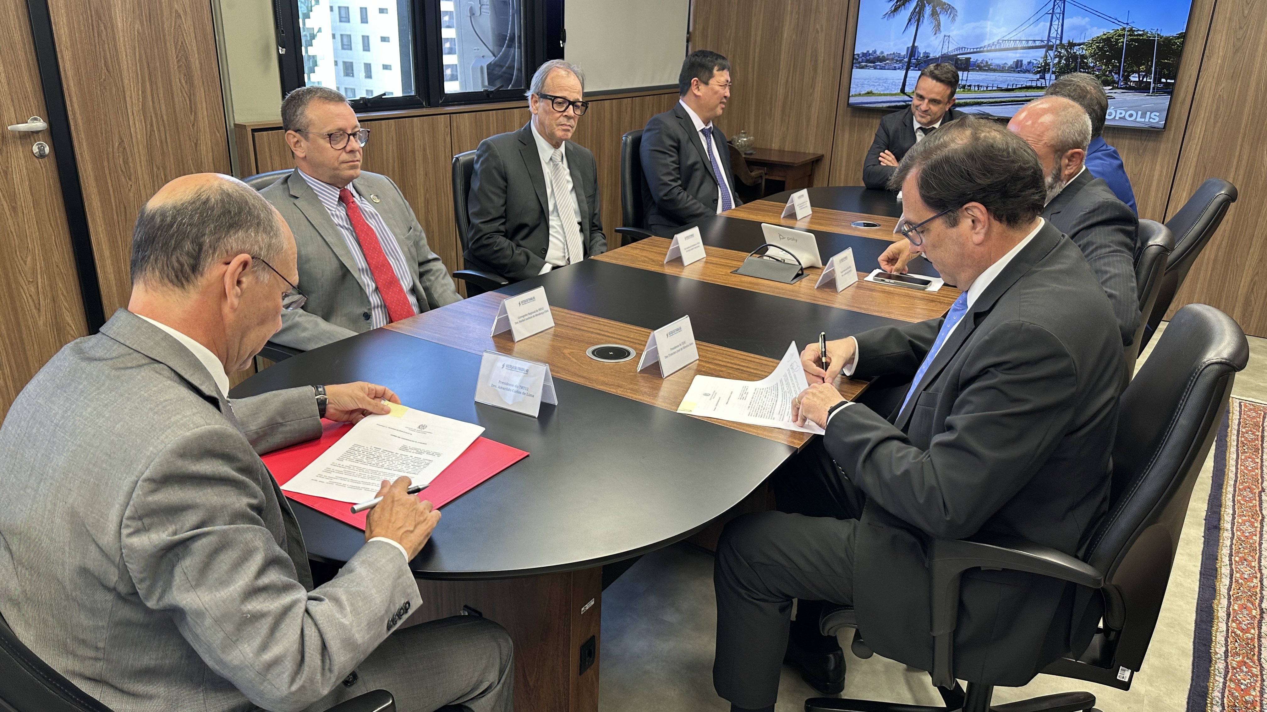 Oito homens de terno e gravata estão sentados em uma mesa oval de reunião. Dois homens estão assinando documentos.