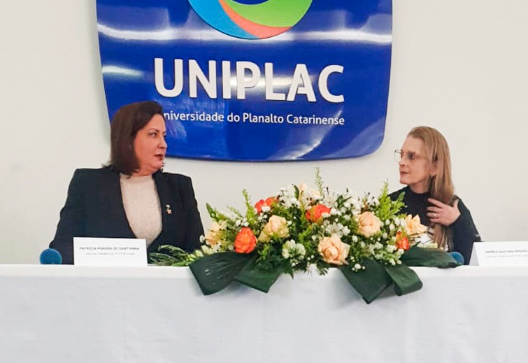 Foto mostra duas mulheres sentadas à frente de uma mesa, com toalha branca e microfones próximos. Entre elas tem um arranjo de flores e, atrás, um banner de lona com o texto UNIPLAC