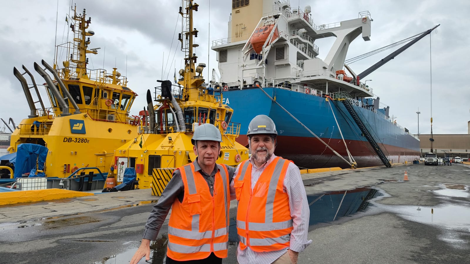 Foto mostra dois homens à frente de um navio cargueiro, em um porto. Eles vestem coletes na cor laranja e capacetes cinzas