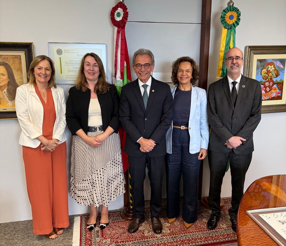 Três mulheres vestidas formalmente e dois homens de terno escuro em pé, lado a lado, sorridentes