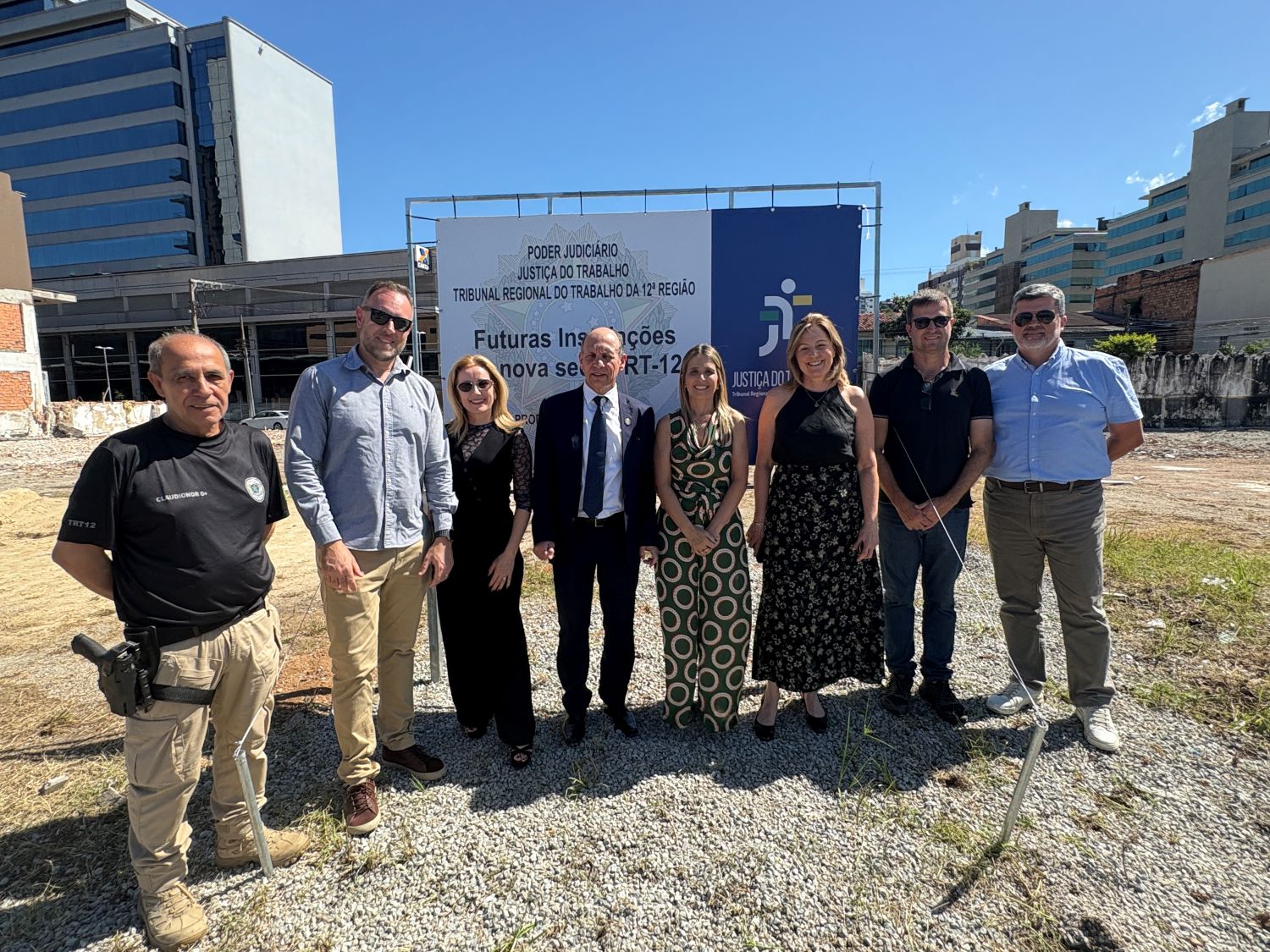Grupo de cinco homens e três mulheres posam em frente a uma placa em um terreno baldio