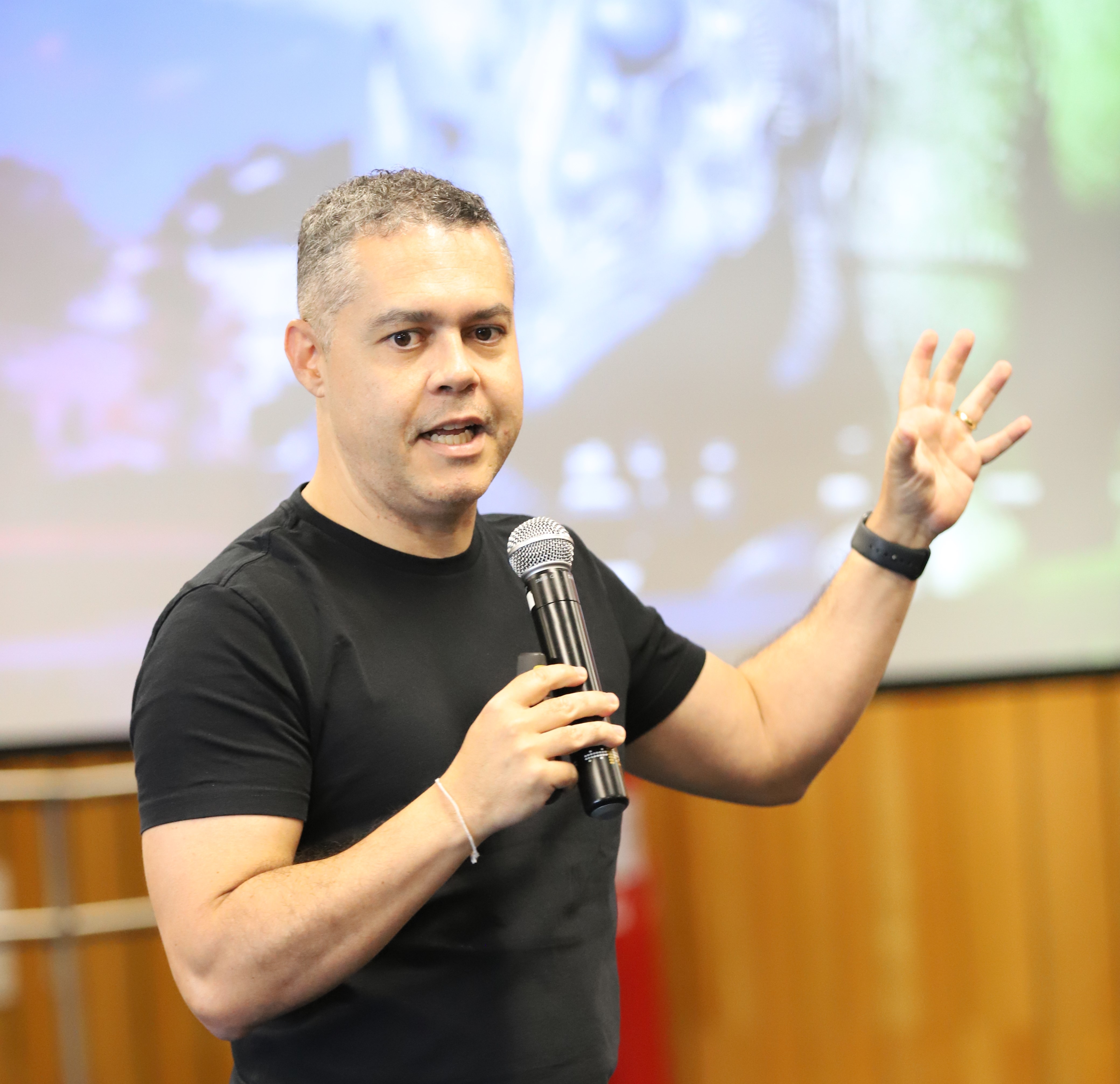 Homem grisalho, de cabelo curto, vestindo camiseta e dando uma palestra. Ao fundo, um painel desfocado em tom verde e lilás