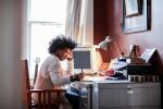 Mulher negra trabalhando em uma mesa, com computador, livros e outros objetos, aparentando estar dentro de um quarto
