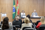 Foto de um auditório com pessoas sentadas, assistindo uma palestra. Na frente do palco uma mulher de cabelo grisalhos e óculos fala no microfone.