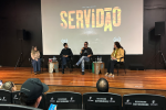 Quatro pessoas estão sentadas em um palco, sendo dois homens e duas mulheres, com uma tela ao fundo na qual está projetado o título do documentário: Servidão.