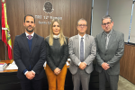 Quatro pessoas posam lado a lado em frente ao letreiro “TRT 12ª Região – Santa Catarina”, dentro de um gabinete. À esquerda, bandeira de Santa Catarina. Todos estão com trajes formais.