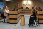 Foto de uma mulher de cadeiras de rodas dando uma palestra. No fundo um painel escrito Tribunal Regional do Trabalho da 12ª Região.