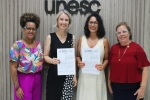 Quatro mulheres estão em pé, posando e sorrindo para a foto, segurando um contrato