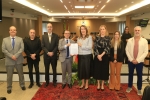 Cinco homens e três mulheres em pé, em uma sala de sessões de julgamento, posam para a foto. Uma mulher e um homem no centro exibem um documento.