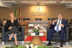 Home de terno escuro fala ao microfone sentado em uma poltrona, na sala de sessões do Pleno do TRT-SC. Ao lado dele, em outra poltrona, uma mulher loira sorridente, com roupa preta