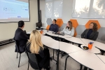 Grupo de três pessoas e dois homens sentados à mesa observando um telão. Uma das mulheres mexe no teclado e esboça um leve sorriso