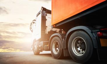 Foto da lateral de um caminhão baú estacionado no asfalto. Ao fundo, ilustração do brilho do sol 