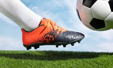 Imagem ilustrativa da visão lateral de um pé com chuteira prestes a chutar uma bola de futebol em um campo gramado