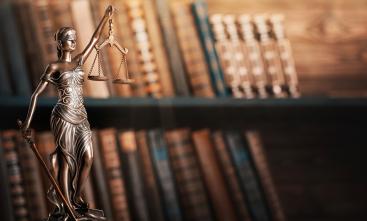 Fotografia mostra escultura da deusa da justiça, Themis, empunhando uma balança em uma das mãos e uma espada na outra, com uma estante de livros ao fundo