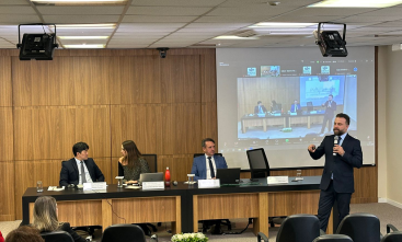 Foto de uma apresentação em um auditório. Um homem branco de cabelo curto, barba e bigode escuro, vestindo terno preto, fala levando um microfone à boca. Atrás dele está uma mesa onde se encontram uma mulher e dois homens sentados e um projetor que transmite uma reunião virtual com enquadramento similar ao da foto.