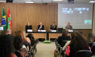 Foto de pessoas sentadas viradas para uma mesa onde estão quatro pessoas. O ambiente é um auditório, à esquerda estão as bandeiras do Brasil e de Santa Catarina e à direita uma reunião virtual sendo projetada