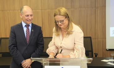 Juíza Ana Rick, uma mulher branca, de cabelos loiros, assina um documento sobre um púlpito enquanto o presidente do TRT-SC, um homem branco, de olhos azuis, observa sorridente.