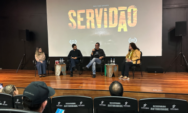 Quatro pessoas estão sentadas em um palco, sendo dois homens e duas mulheres, com uma tela ao fundo na qual está projetado o título do documentário: Servidão.