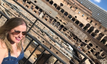 Uma mulher loira com óculos escuros sorri para a foto. Ao fundo o Coliseu, em Roma